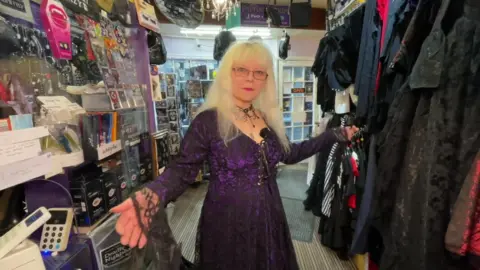 Une femme avec des cheveux blancs et des lunettes, portant une robe noire et violette en dentelle, entourée de vêtements gothiques dans un magasin.