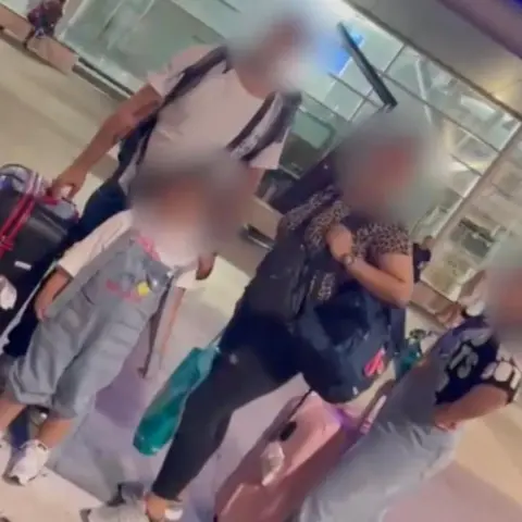 Une famille de migrants pose devant un aéroport. Leurs visages sont floutés