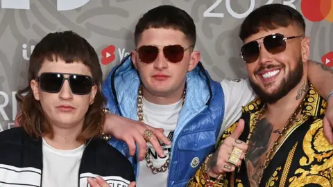 Les membres du Bad Boy Chiller Crew lors des Brit Awards en 2023, vêtus de tenues colorées avec des chaînes en or et des lunettes de soleil.