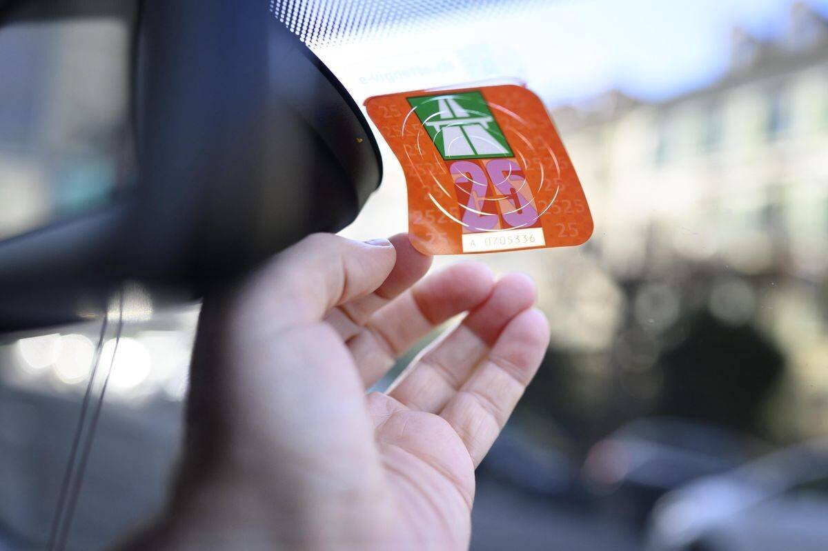 Un homme colle une vignette autoroutière pour l'année 2025 sur le pare-brise d'une voiture, photographié à Bern. (KEYSTONE/Christian Beutler)