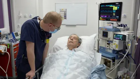 Un homme allongé dans un lit d'hôpital avec une infirmière qui lui parle.