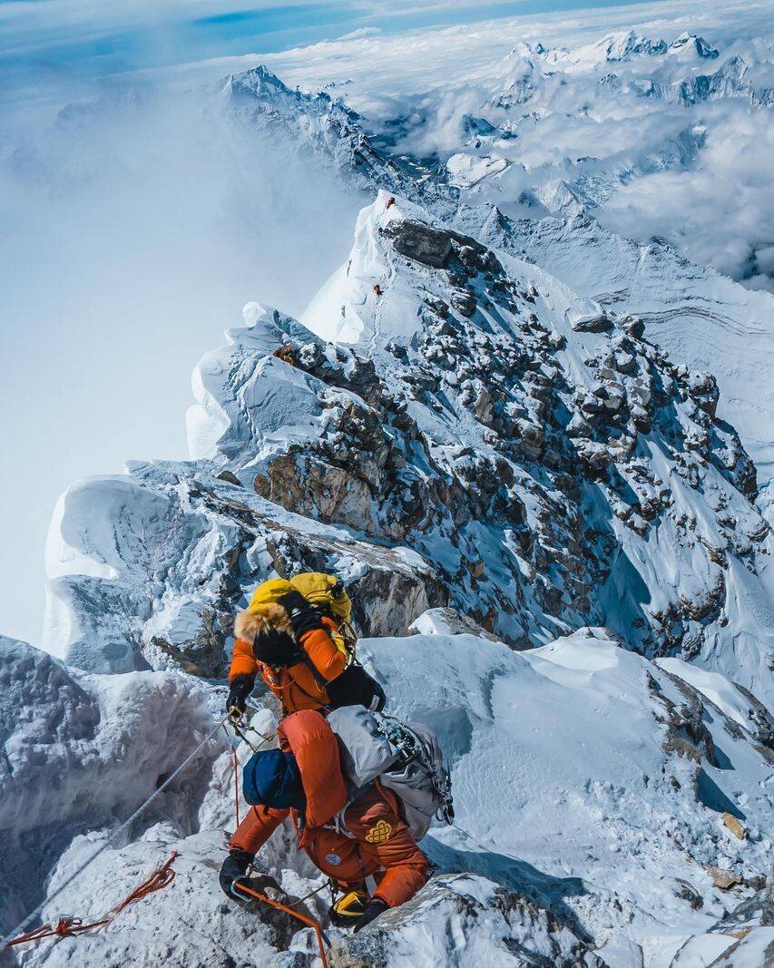 Bergsteiger in orangefarbenen Anzügen erklimmen einen schneebedeckten Grat in einer hochalpinen Landschaft.