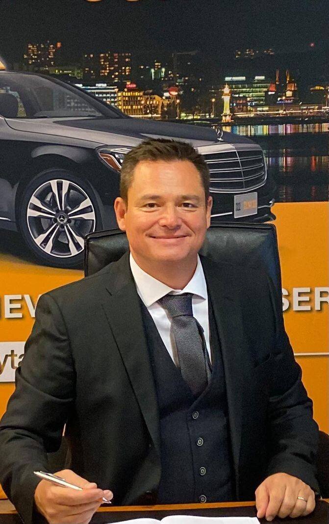 Cédric Bouchard, patron de Taxiphone, en costume assis à un bureau, tenant un stylo, avec une affiche de voiture et vue nocturne en arrière-plan.