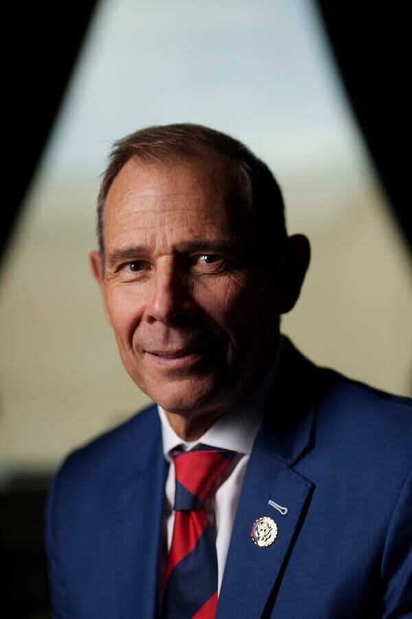 Portrait de John Curtis portant un costume bleu et une cravate rayée rouge.