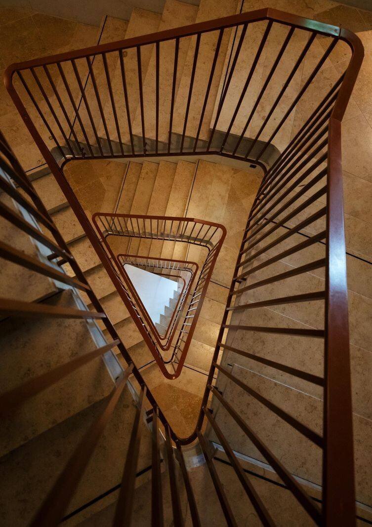 Treppenhaus des Lighthouse-Palliativzentrums in Zürich: Triangularer Treppenhausbereich mit braunem Geländer und Marmorstufen, die sich in die Tiefe erstrecken.