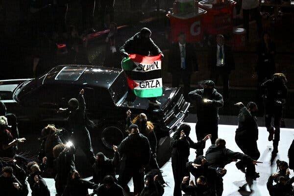 Une personne debout sur un véhicule avec un drapeau portant les mots 'Soudan' et 'Gaza'.