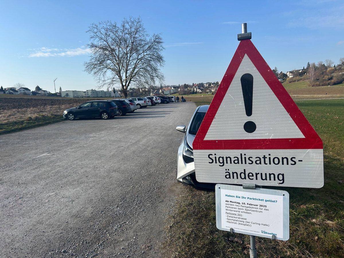 Ein Parkplatz an einem sonnigen Tag mit einem Warnschild vorne, das auf eine Signalisationsänderung hinweist.