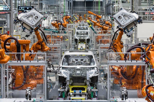 Car bodies on an assembly line, with orange machines on both sides. The two machines in front are holding car doors.