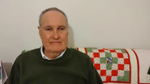 David Evans, assis sur un canapé, montre une expression de colère.