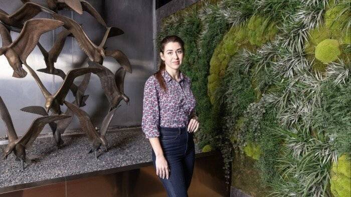 Une femme en chemise à fleurs et jeans se tient à côté d'un mur de plantes vertes et d'une sculpture d'oiseau en métal dans un cadre intérieur