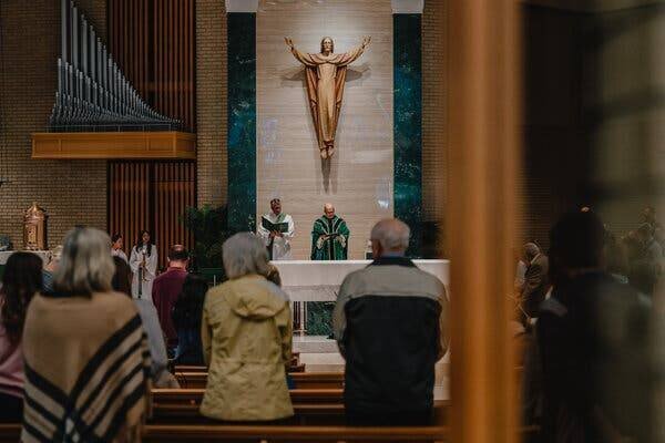 Des personnes se tiennent parmi les bancs d'une église pendant que deux prêtres dirigent la messe.