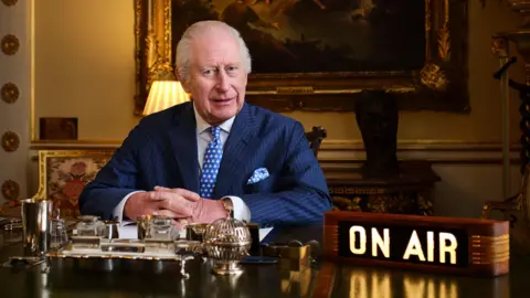Le Roi Charles assis derrière un bureau avec un panneau 'on air' à ses côtés