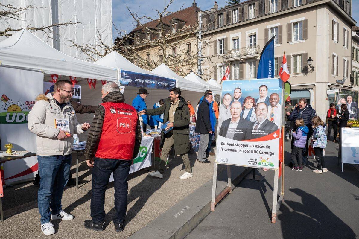 Stands des élections municipales carougeoises 2025 à la place du Marché à Carouge, avec des affiches de candidats et des gens discutant.
