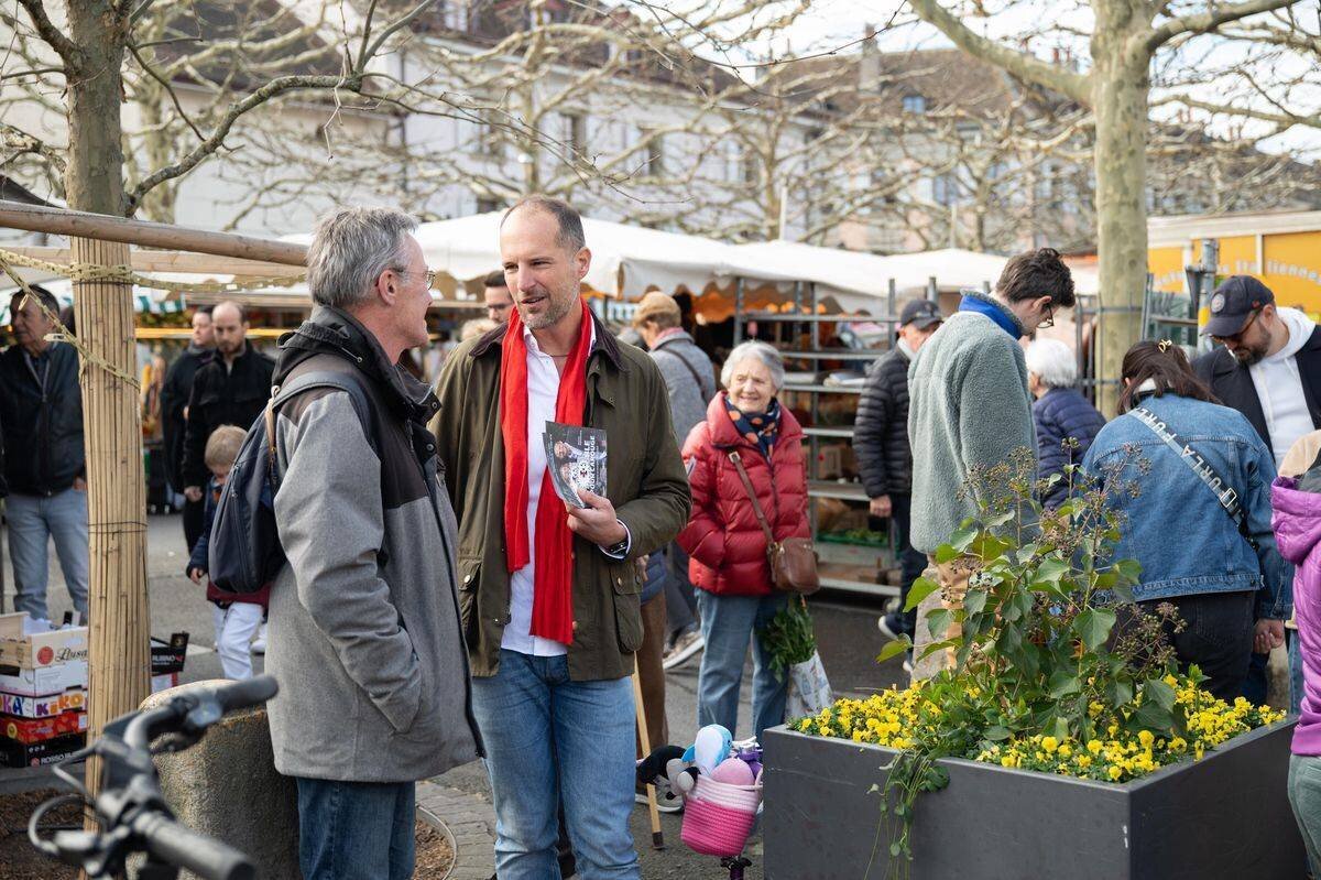 Stand pour les élections municipales 2025 à Carouge, place du Marché, avec des personnes discutant et passant parmi les stands.