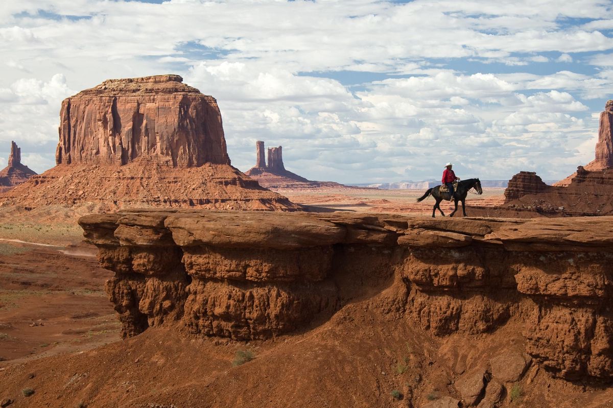 Paysage désertique avec une personne à cheval sur une falaise rocheuse, formation rocheuse emblématique à l’arrière-plan.