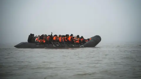 Un canot en caoutchouc noir rempli de passagers, la plupart portant des gilets de sauvetage orange, flotte dans le brouillard sur la Manche. Une personne lève la main.