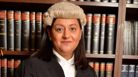 La juge principale Aisha Answar devant une bibliothèque remplie de livres de droit, souriante.