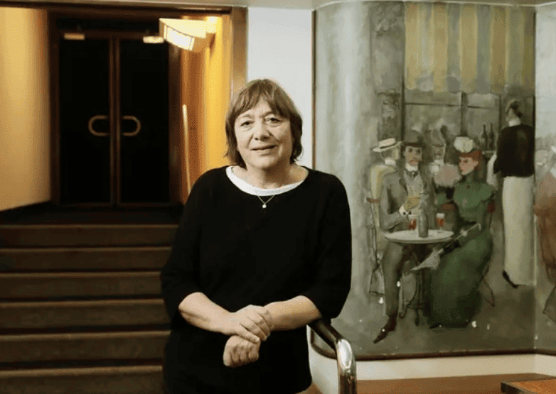Une femme souriante, vêtue de noir, se tient debout devant un tableau représentant une scène de café parisien.