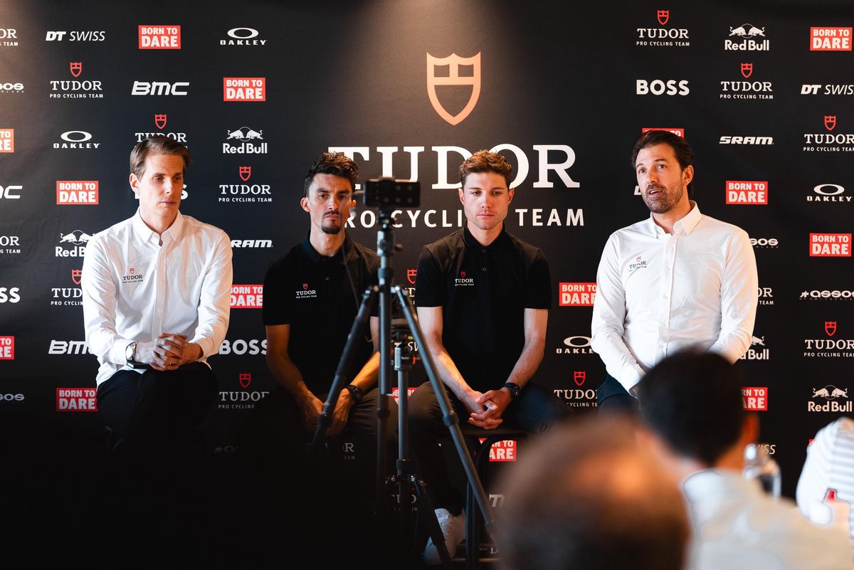 Vier Männer in einem Interview mit dem Tudor Pro Cycling Team, sitzend vor einer schwarzen Wand mit Sponsorlogos wie Tudor, Red Bull und Boss.