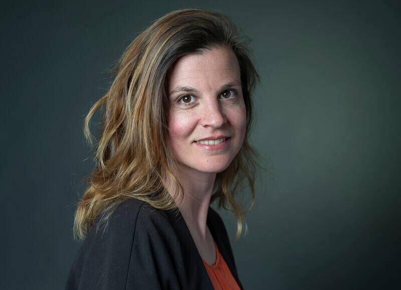 Femme souriante aux cheveux châtain clair, portant un haut rouge et une veste noire, posant devant un fond vert.