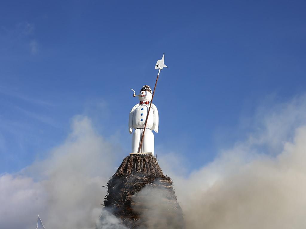 Der Böögg, eine Schneefigur, sitzt auf einem Holzhaufen, umgeben von Rauch, unter blauem Himmel beim Zürcher Frühlingsfest.