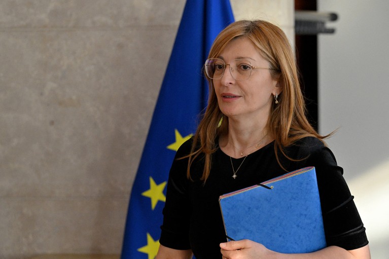 Ekaterina Zaharieva, commissaire européenne pour les start-ups, la recherche et l'innovation, tenant un dossier bleu devant un drapeau de l'UE.