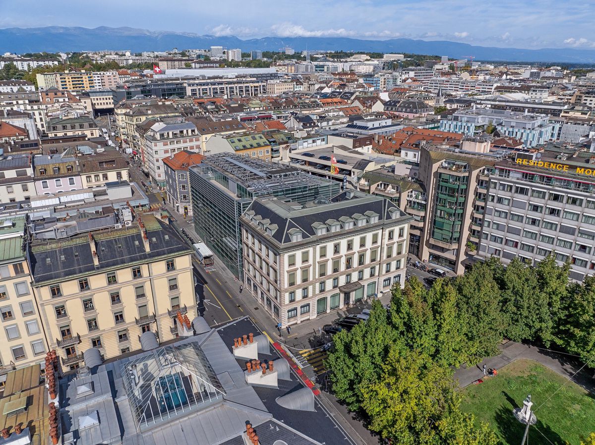 Vue aérienne de Genève, montrant des bâtiments du centre-ville, la verdure d’un parc et des montagnes en arrière-plan.