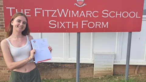 Emma, souriante tenant fièrement ses résultats devant son établissement scolaire