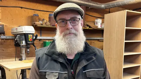 Mark Lucas dans un atelier de menuiserie entouré d’outils et d’étagères en bois