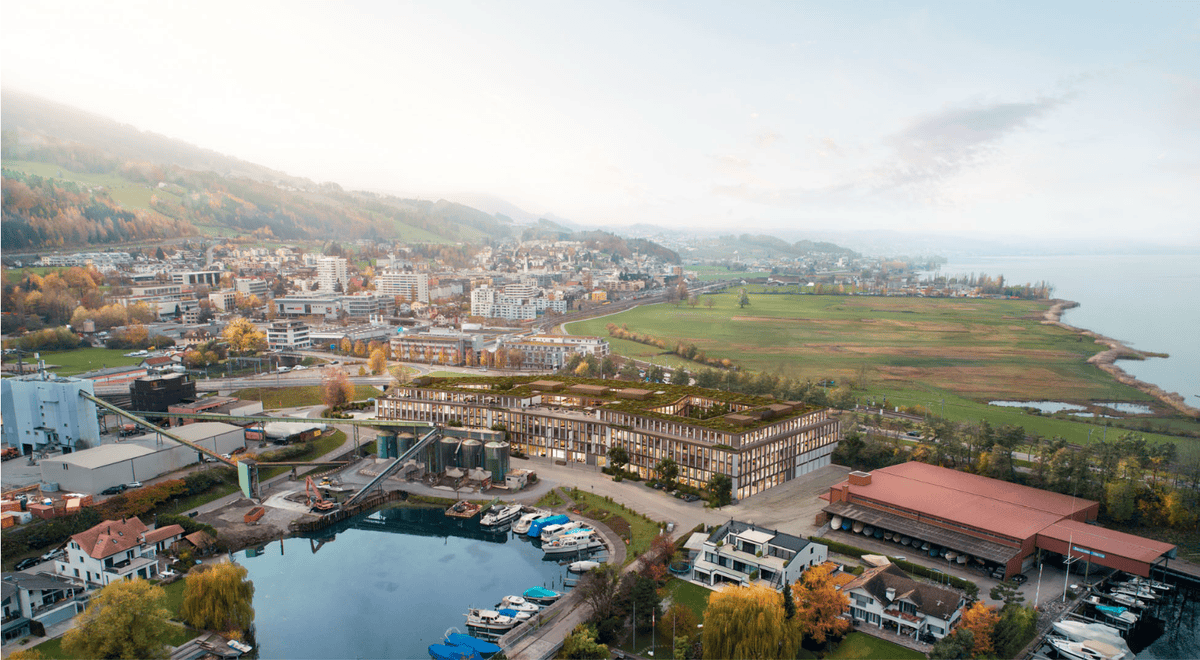Vue aérienne d’un paysage urbain avec bâtiments, espaces verts et lac en arrière-plan sous un ciel partiellement nuageux.