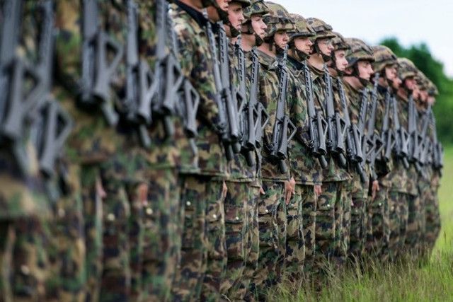 Soldats en uniforme de camouflage alignés en rangée, portant des fusils, sur l’herbe verte.