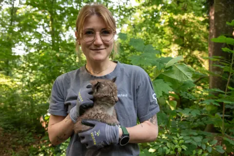 Dina Gebhardt tenant un bébé lynx et souriante devant une forêt
