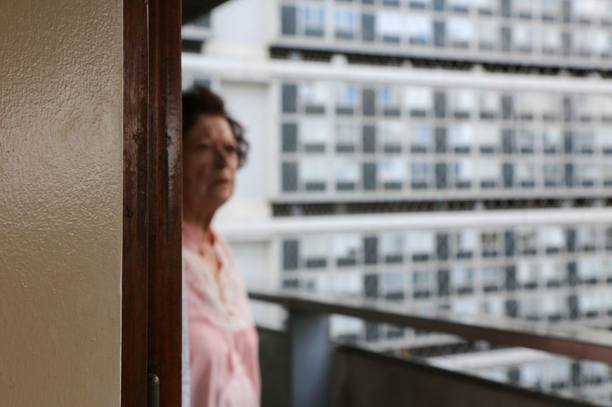 Une femme floue debout sur un balcon avec une grande façade d’immeuble en arrière-plan.