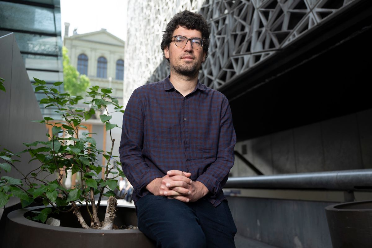 Hadrien Buclin, député d’Ensemble à Gauche, assis à Lausanne, le 02 mai 2025, cherchant à déposer une requête pour une commission d’enquête parlementaire sur le bouclier fiscal. En arrière-plan, un bâtiment moderne. ©Florian Cella / Tamedia