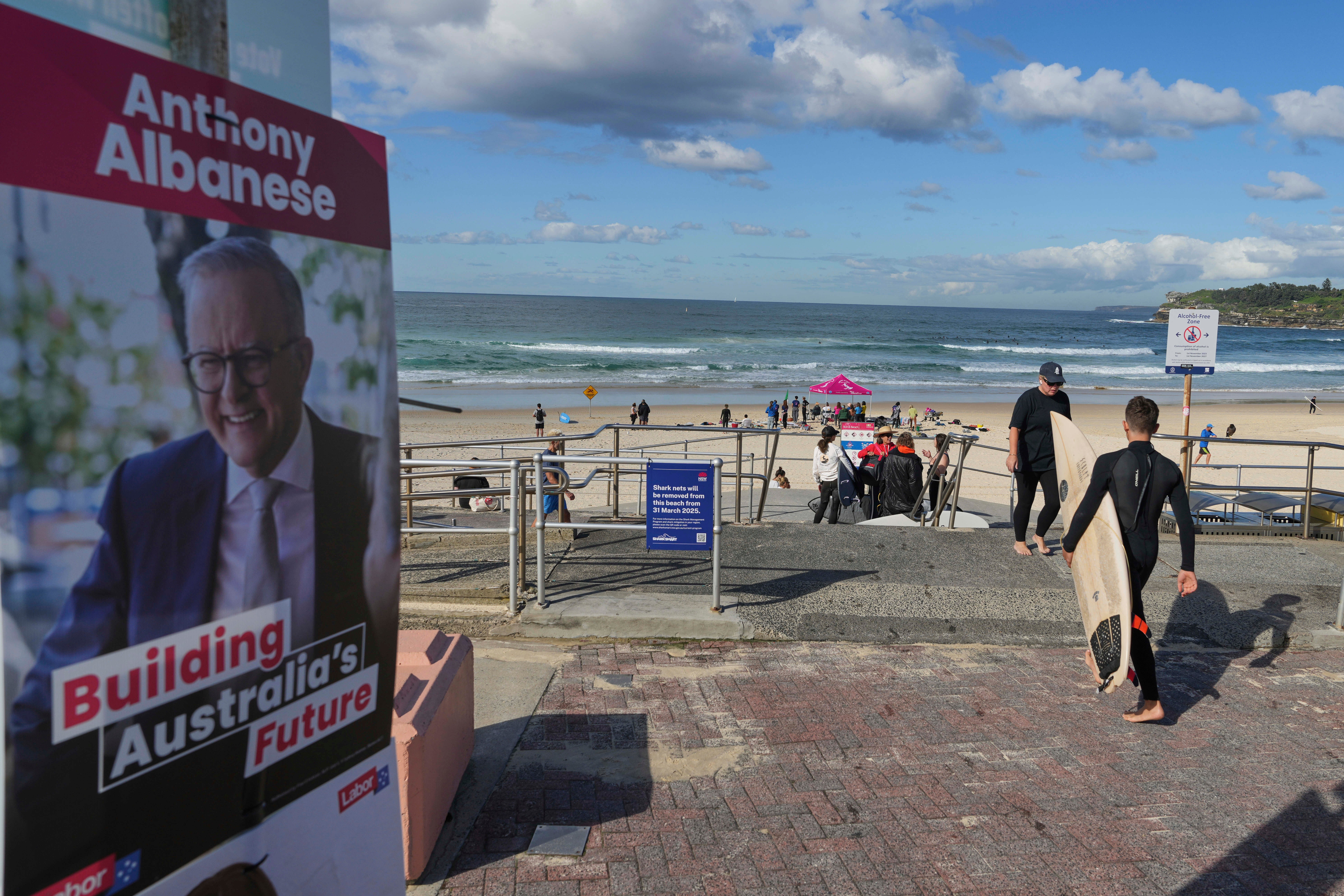 Un surfeur porte sa planche devant une photo du Premier ministre Anthony Albanese près d'un bureau de vote à Bondi Beach, Sydney, samedi 3 mai 2025