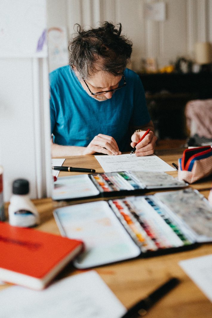 Homme concentré, vêtu d’un t-shirt bleu, dessinant sur un bureau encombré d’aquarelles et de fournitures artistiques.