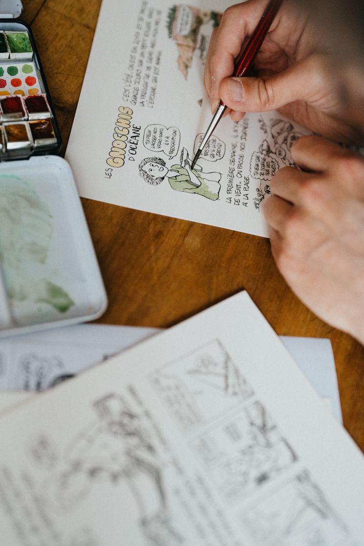 Personne dessinant des croquis sur du papier avec un pinceau, entourée de palettes de couleurs.