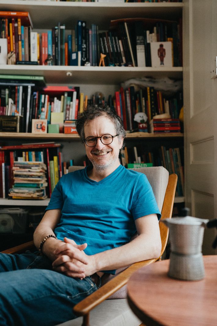 Un homme souriant portant un t-shirt bleu est assis sur une chaise devant une bibliothèque bien garnie.