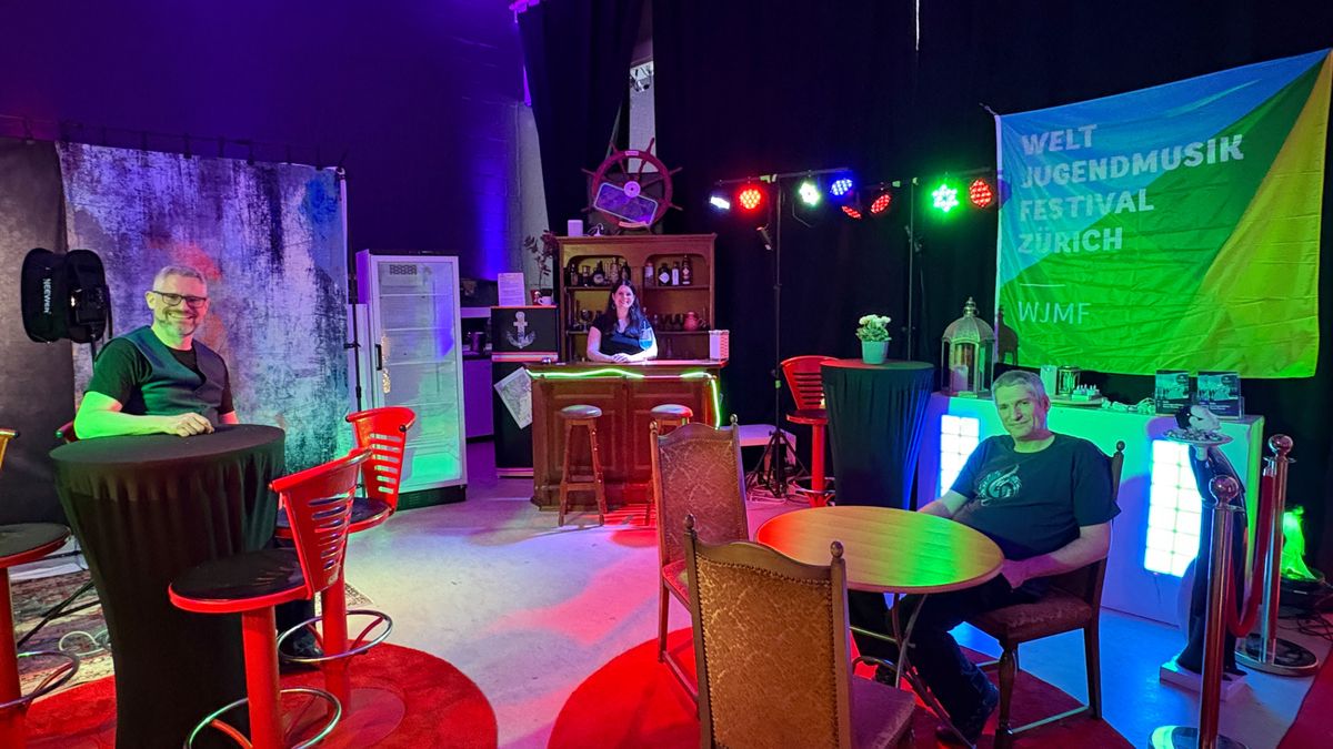 Trois personnes assises autour d'une table dans un espace avec un bar et une bannière du festival mondial de musique jeunesse de Zurich
