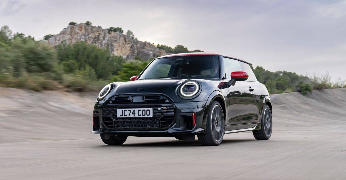 Schwarzer Mini Cooper mit rotem Dach fährt auf einer kurvigen Strasse vor bewaldeter Hügelkulisse an einem bewölkten Tag.