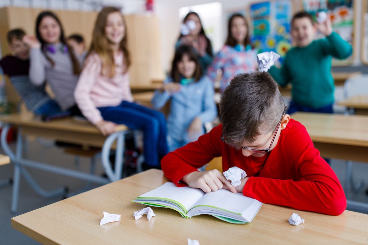 Un garçon se fait intimider à l’école, recroquevillé sur un bureau, entouré d’autres enfants se moquant de lui.
