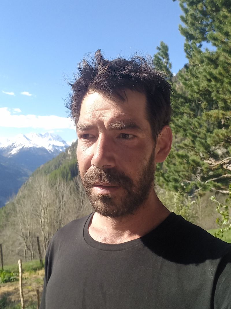 Homme barbu en plein air, fond de montagnes enneigées et arbres verts sous un ciel bleu clair.
