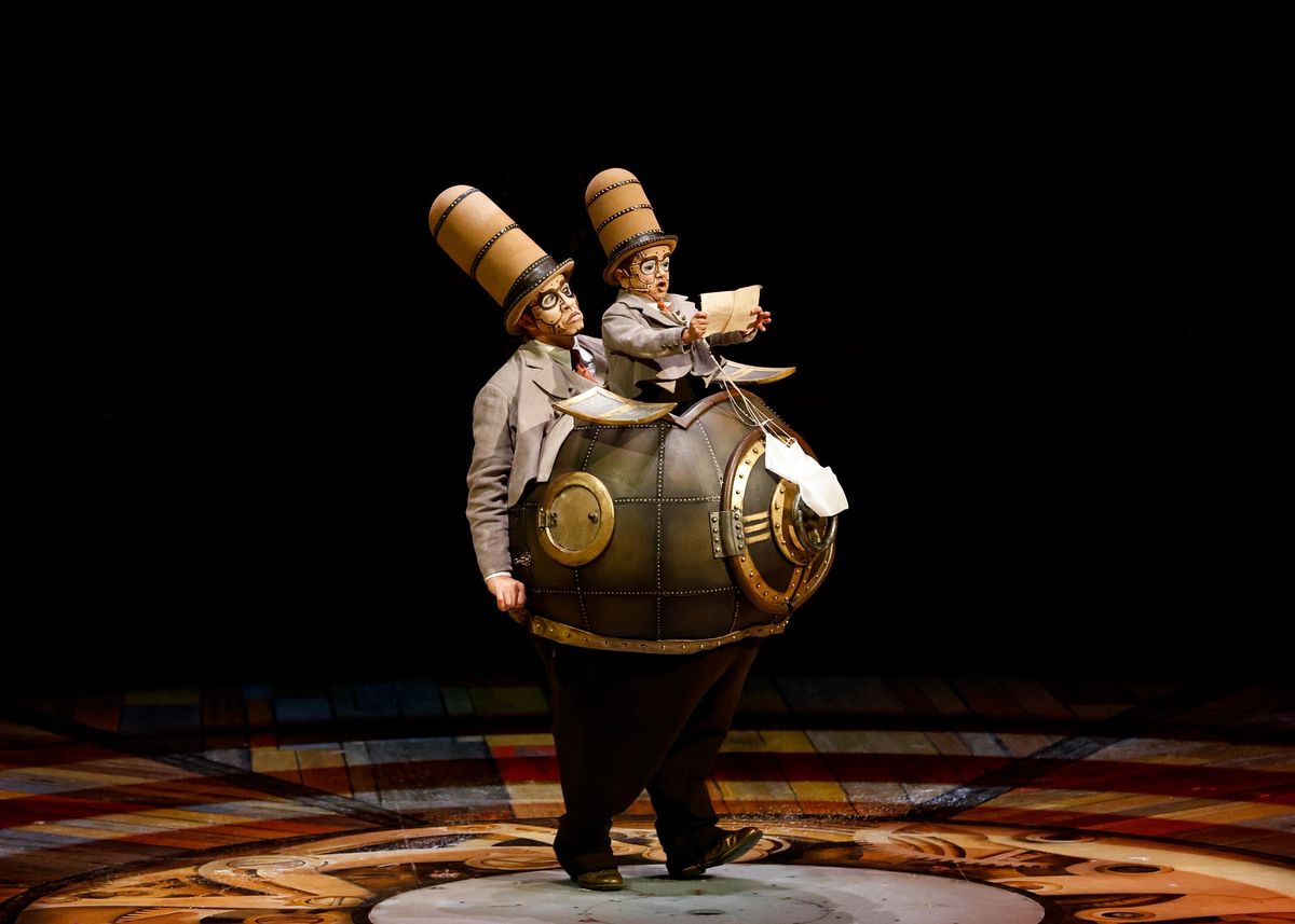 Deux artistes en costumes extravagants sur scène, portant de grands chapeaux cylindriques et un costume en forme de machine.