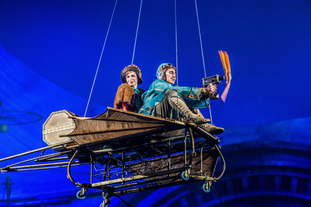 Deux artistes en costume montent sur un appareil volant lors d’une performance de cirque sous un chapiteau bleu.