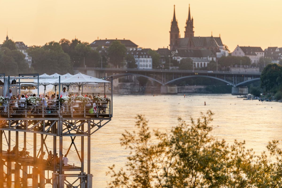 Vue sur le Rhin Bleu et la cathédrale de Bâle en été, avec une terrasse animée surplombant la rivière.