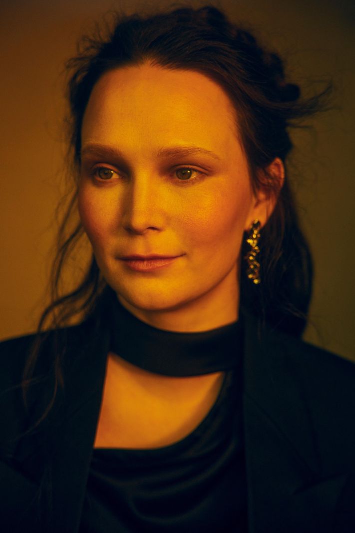 Portrait de Zoë Më, représentante de la Suisse à l’Eurovision 2025, avec un fond jaune et une tenue élégante.