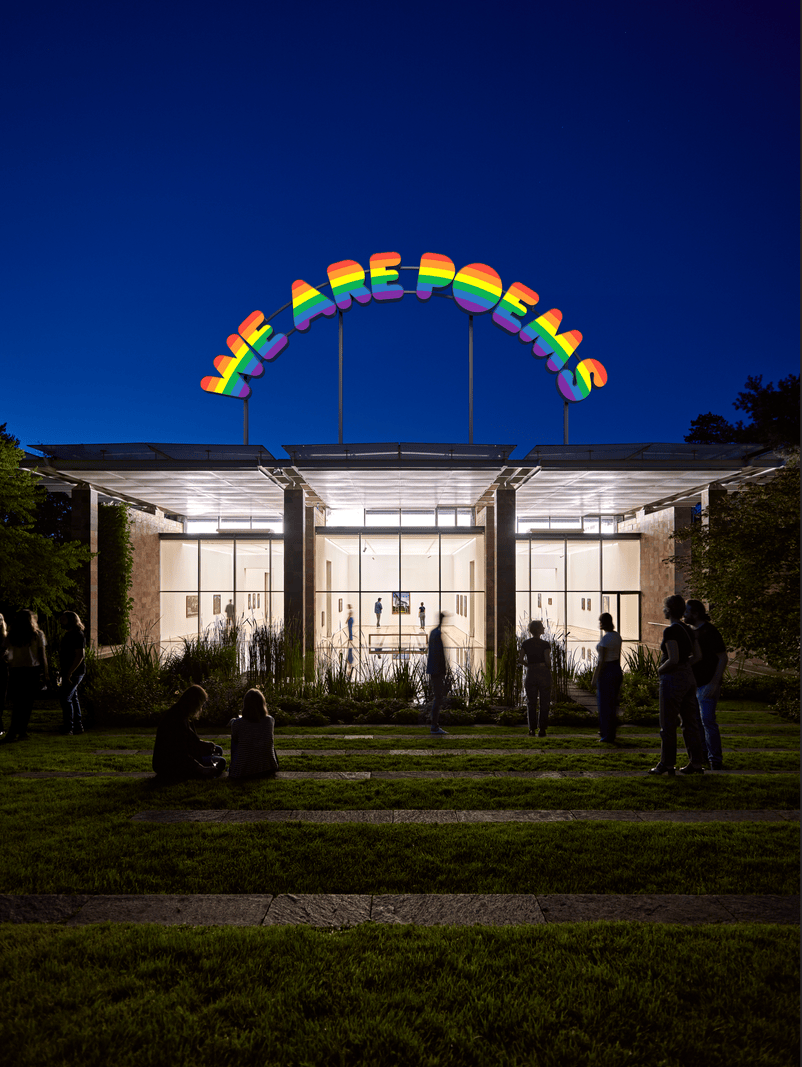 Bâtiment moderne éclairé la nuit avec une enseigne colorée ’WE ARE POEMS’, entouré de personnes debout et assises à l’extérieur.