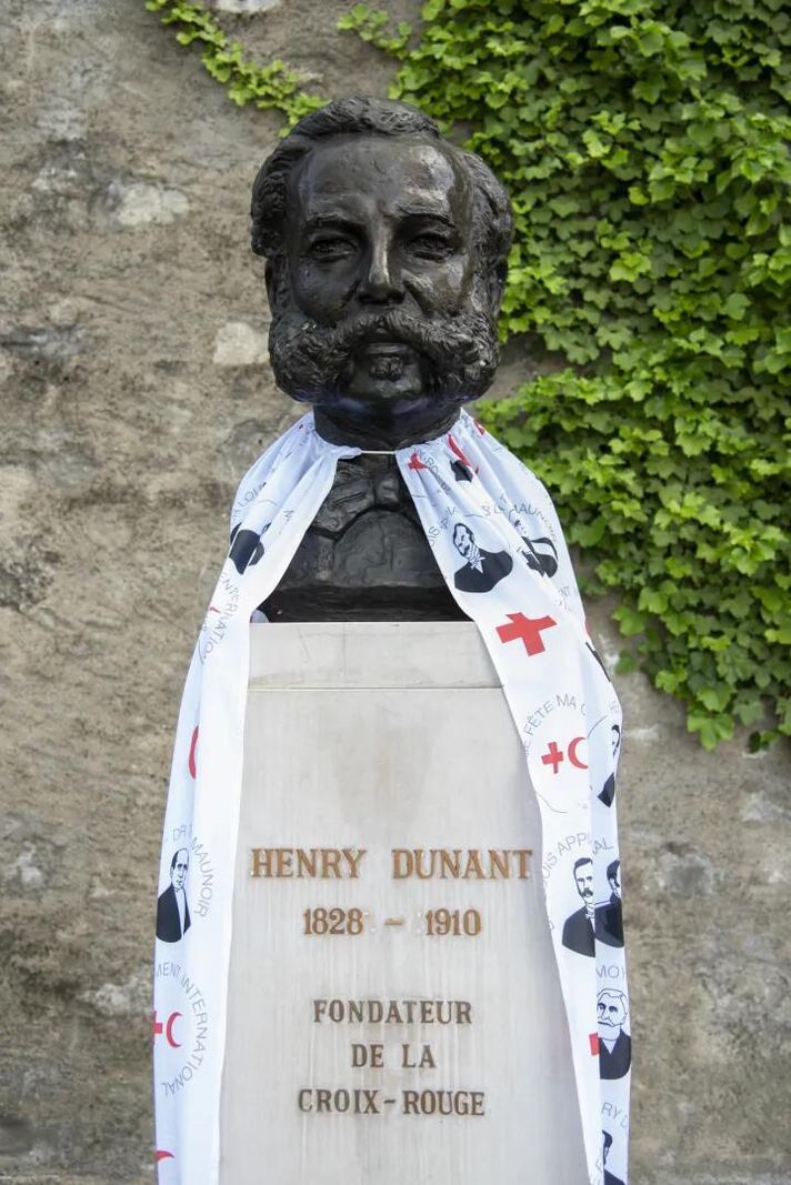 Buste d'Henry Dunant recouvert d’une écharpe illustrée, fondateur de la Croix-Rouge, avec un mur de pierre et de la verdure en arrière-plan.
