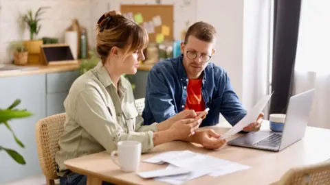 Couple examine ses finances autour de la table de la cuisine avec des factures, un ordinateur portable et une tasse devant eux.