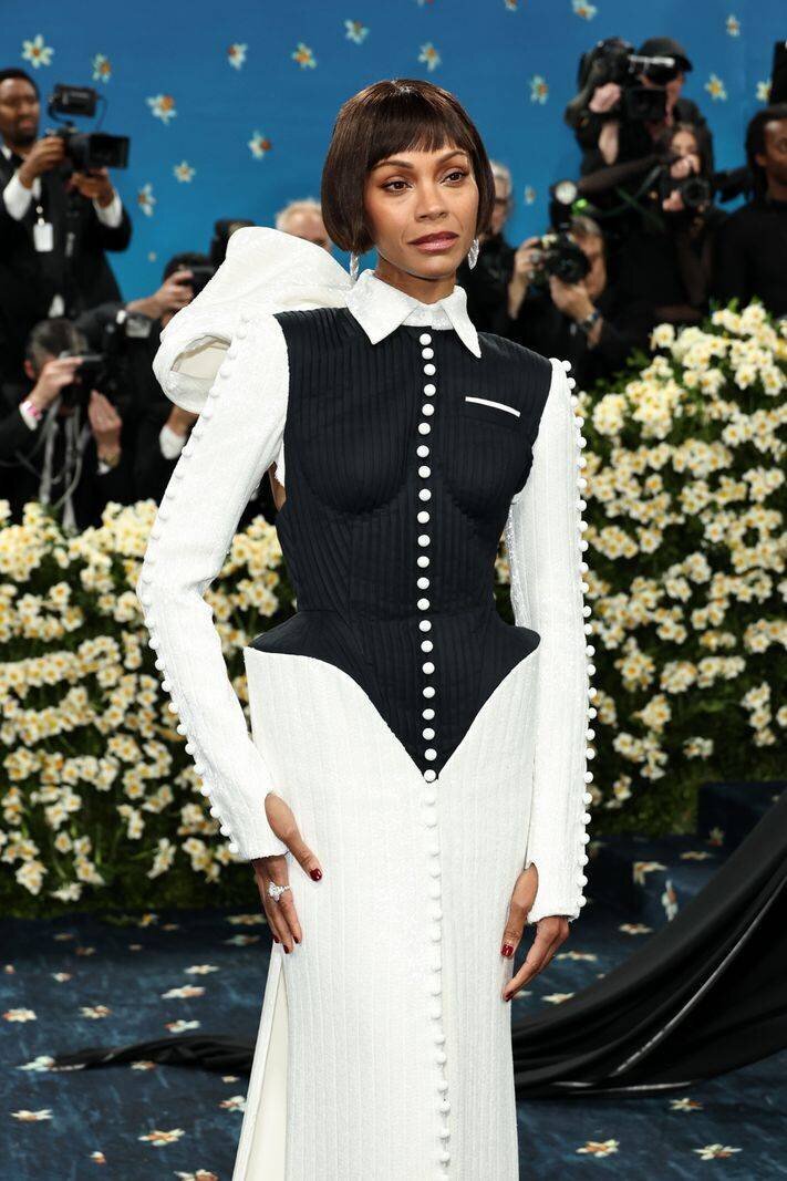 Zoe Saldana au Met Gala 2025, portant une robe noir et blanc à boutons, entourée de fleurs blanches et de photographes.